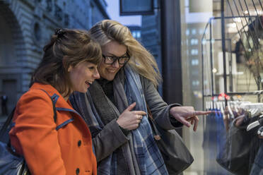 Smiling young woman pointing at glass while doing window shopping with female friend - WPEF04833