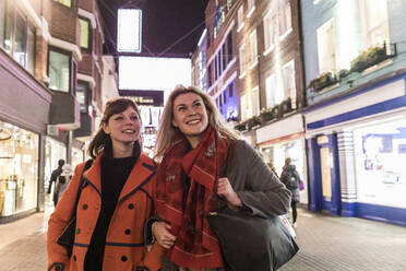 Lächelnde Freundinnen in Wintermänteln schauen weg, während sie zusammen in der nächtlichen Stadt spazieren gehen - WPEF04837