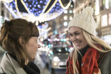 Smiling blond woman wearing knit hat looking at female friend while talking on Christmas - WPEF04843