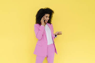 Lächelnde weibliche Fachkraft gestikuliert, während sie mit einem Handy vor einem gelben Hintergrund spricht - KNSF08701