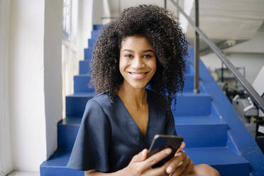Lächelnde Unternehmerin mit Mobiltelefon auf den Stufen im Büro sitzend - KNSF08711