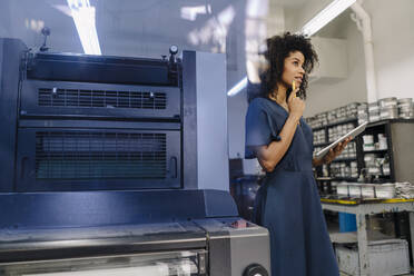 Nachdenkliche Geschäftsfrau mit digitalem Tablet, die wegschaut, während sie an einer Maschine in der Industrie steht - KNSF08749