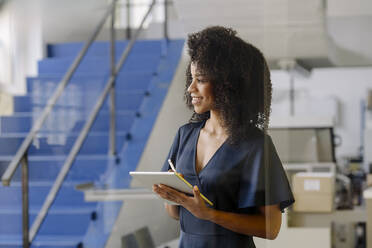 Lächelnde weibliche Fachkraft, die ein digitales Tablet hält, während sie an einer Treppe in der Industrie steht - KNSF08754