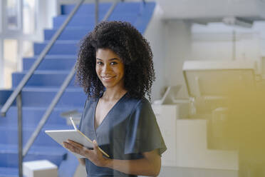 Lächelnde Afro-Fachfrau mit digitalem Tablet, die an einer Treppe in der Industrie steht - KNSF08755