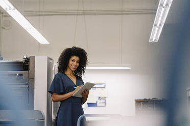 Smiling Afro female entrepreneur looking away while working on digital tablet at industry - KNSF08760