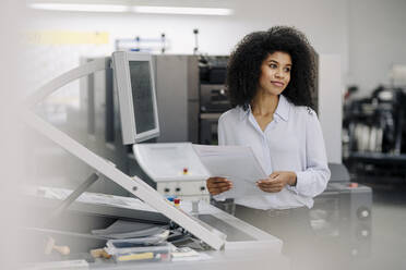 Afro-Geschäftsfrau beim Betrachten von Dokumenten an einer Maschine in der Industrie - KNSF08776