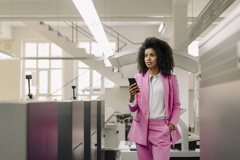 Weibliche Fachkraft mit Mobiltelefon schaut weg, während sie inmitten von Maschinen in der Industrie steht - KNSF08789