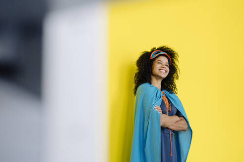 Lächelnde Frau im Superheldenkostüm schaut weg, während sie vor einer gelben Wand steht - KNSF08809