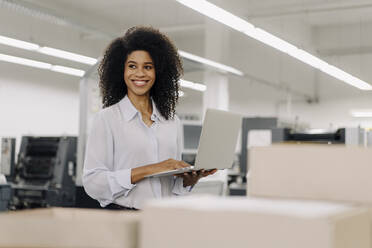 Lächelnde weibliche Fachkraft, die wegschaut, während sie einen Laptop in der Industrie hält - KNSF08832