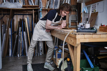 Male store owner wearing apron painting picture frame while leaning on table - ASGF00500