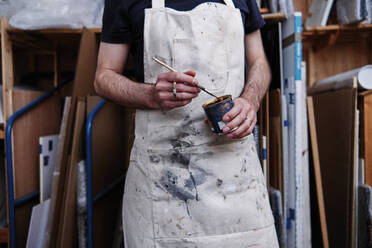 Male frame maker wearing apron holding paint brush and can while standing in workshop - ASGF00502