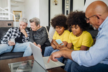 Twin boys with grandfather using laptop while young man using smart phone sitting by great grandmother in living room - MEUF03117