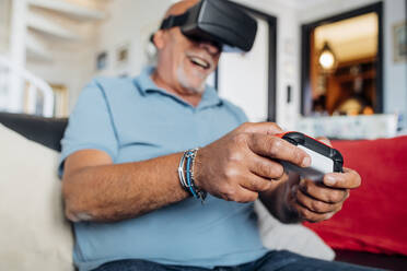 Cheerful senior man wearing virtual reality simulator playing video game at home - MEUF03125