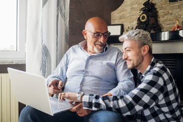 Lächelnder Vater und Sohn teilen sich einen Laptop, während sie zusammen zu Hause sitzen - MEUF03128