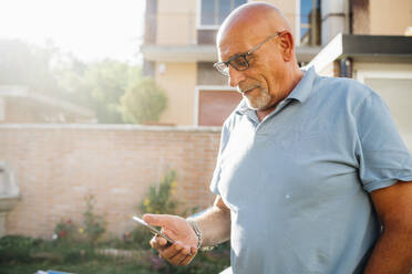 Senior man wearing eyeglasses using smart phone while standing at backyard during sunny day - MEUF03130