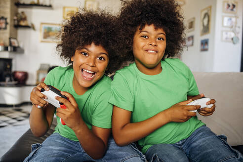 Cheerful twin boys with curly hair playing video games sitting on sofa in living room at home - MEUF03133