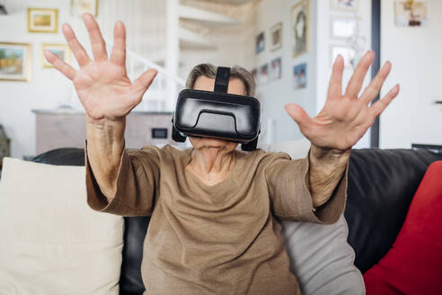 Ältere Frau trägt einen Virtual-Reality-Simulator und gestikuliert, während sie zu Hause auf dem Sofa im Wohnzimmer sitzt - MEUF03135