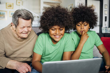 Lächelnder Junge, der einen Laptop benutzt, während er neben seiner Urgroßmutter und seinem Bruder im Wohnzimmer sitzt - MEUF03136