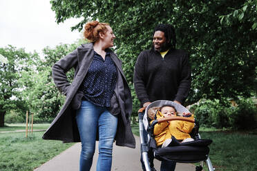 Vater spricht mit Mutter, während er seinen Sohn im Kinderwagen durch den Park schiebt - ASGF00520