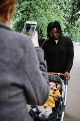 Vater hält Baby im Kinderwagen, während Frau im Park fotografiert - ASGF00525