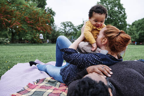 Mutter küsst ihren Sohn und lehnt sich an einen Mann, der auf einer Picknickdecke im Park liegt - ASGF00541