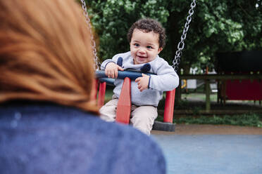 Lächelnder Junge schaut Frau an, während er auf einer Schaukel auf dem Spielplatz sitzt - ASGF00570