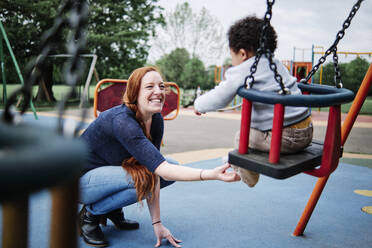 Mother tickling son sitting on swing in playground at park - ASGF00571