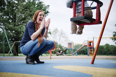 Glückliche Mutter klatscht, während sie in der Nähe ihres Sohnes, der auf einer Schaukel auf dem Spielplatz sitzt, hockt - ASGF00572