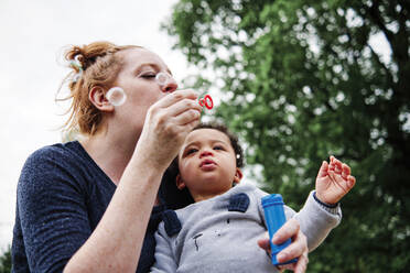 Mother blowing bubbles with son at park - ASGF00574