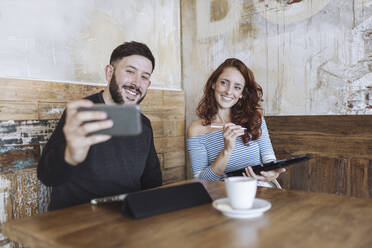 Mann macht Selfie mit einer Kollegin, die ein Grafiktablett in einem Café hält - MTBF01026