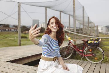 Frau, die ein Selfie mit ihrem Smartphone macht, während sie auf dem Pier sitzt - MTBF01030