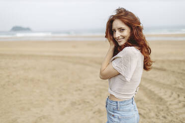 Lächelnde rothaarige Frau am Strand stehend - MTBF01040