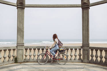 Frau schaut auf das Meer, während sie auf einem Fahrrad in der Nähe einer Stützmauer sitzt - MTBF01044