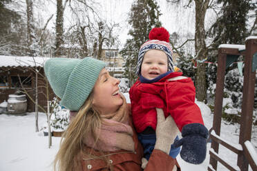 Cheerful mother carrying cute son while standing at backyard during winter - FVDF00227