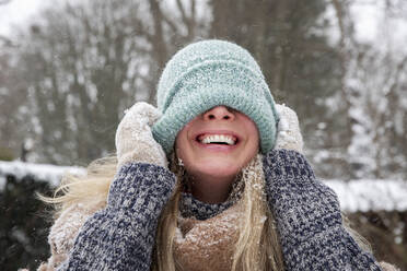 Cheerful woman covering face with knit hat during winter - FVDF00234