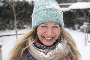 Cheerful blond woman wearing knit hat and scarf standing at backyard - FVDF00241