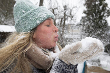 Blond woman with eyes closed blowing snow while standing at backyard during winter - FVDF00245