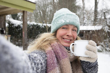 Lächelnde reife Frau, die eine Kaffeetasse hält, während sie im Hinterhof im Schnee steht - FVDF00250