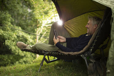 Man in camping lounge chair fishing and using digital tablet stock