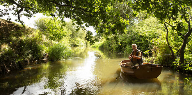 Mann beim Fliegenfischen vom Boot aus auf einem ruhigen grünen Fluss - CAIF30694