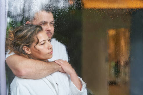 Gelassenes, zärtliches Paar, das sich am regnerischen Fenster umarmt - CAIF30704