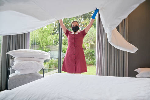 Weibliches Hotelzimmermädchen mit Gesichtsmaske beim Bettenmachen im Hotelzimmer - CAIF30710