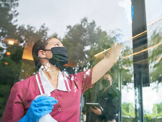 Weibliches Hotelzimmermädchen mit Gesichtsmaske putzt Hotelfenster - CAIF30712