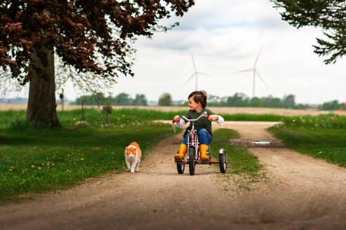 Junge fährt Dreirad in der Einfahrt mit orange-weißer Hauskatze - CAVF94155