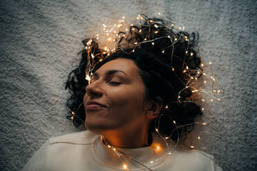 Woman lying down with tangled string lights in her hair, smiling - CAVF94162