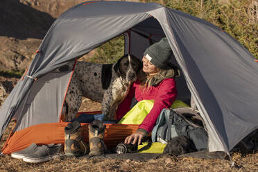 Lächelnde junge Frau mit Hund im Zelt auf einem Berg im Urlaub - CAVF94183