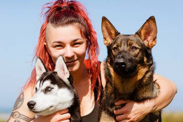 Content female athlete with piercing embracing Siberian Husky and German Shepherd while looking at camera in sunlight - ADSF24705