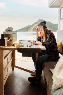 Side view of focused female traveler creating handmade accessories while sitting at wooden table in parked truck at seaside - ADSF24710