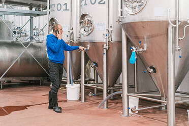 Seitenansicht eines männlichen Unternehmers, der mit einem Handy spricht, vor Edelstahlbehältern auf nassem Boden in einer Bierfabrik - ADSF24720