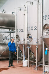 Seitenansicht eines männlichen Unternehmers, der mit einem Handy spricht, vor Edelstahlbehältern auf nassem Boden in einer Bierfabrik - ADSF24721